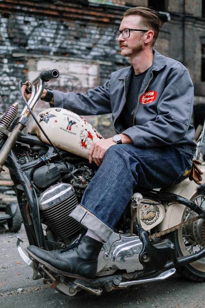Fashionable man on a vintage motorcycle in an urban setting wearing eyeglasses and jeans.