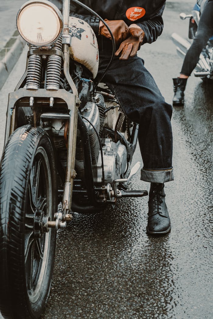 Person with vintage motorcycle on wet street, wearing black boots and denim.